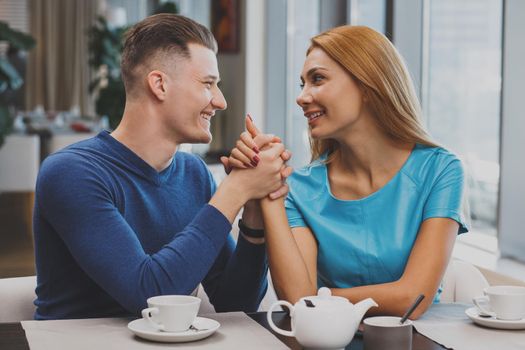 Young happy loving couple holding hands at the restaurant, smiling at each other. Handsome young man holding hands with his girlfriend at the cafe, looking each other in the eyes
