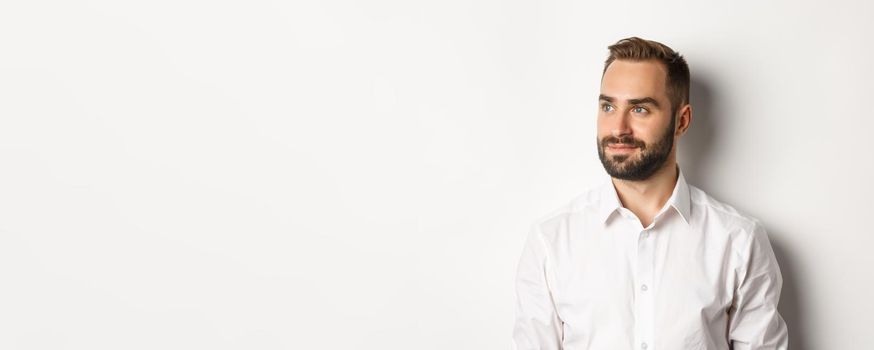 Close-up of confident businessman in white shirt, looking left and smiling satisfied, standing over white background.