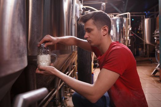 Young handsome man working at his microbrewery, pouring beer from metal tank into glass