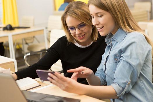 Beautiful young woman showing something on her smart phone to a female friend sitting together in front of the laptop at the office designers creative team working studying friendship mobility carrier