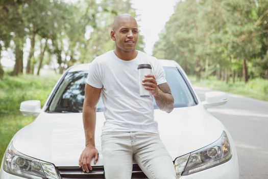 Charming young African man smiling, enjoying drinking coffee on a countryside road, leaning on his car. Attractive man resting from driving automobile during his roadtrip