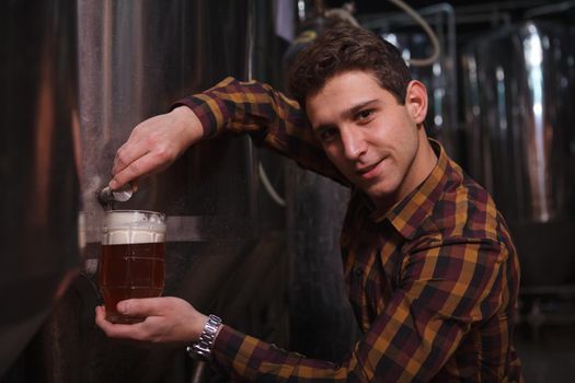 Handsome cheerful brewer looking to the camera while pouring freshly brewed beer from the tank into a mug at his microbrewery