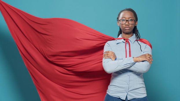 Woman superhero posing with arms crossed in studio, wearing red cape cartoon costume on camera. Feeling confident and powerful as comic character with cloak, flying action concept.