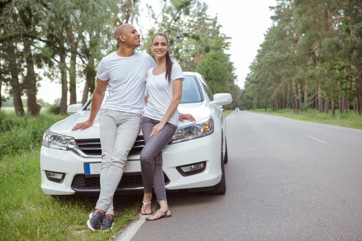 Happy young mixed race couple hugging near their car on the road. Beautiful woman enjoying roadtrip with her handsome boyfriend
