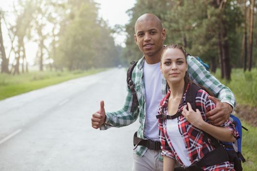 Young travelers couple hitchhiking on the road, copy space. Handsome African man and his beautiful girlfriend catching a car on the side of the road, while backpacking together