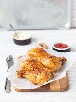 Roasted golden chicken legs drumsticks on baking paper on wooden board served with ketchup and mayonnaise on rustic white wooden table, angle view