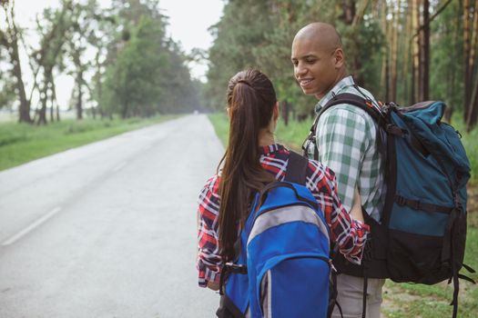 Handsome African man with backpack smiling at his girlfriend while hitchhiking on the countryside road, copy space. Young couple travelling together