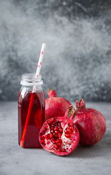 Freshly squeezed red pomegranate healthy juice in bottle with paper straw near whole and split in half fresh organic ripe pomegranate fruit with seed on grey concrete background .