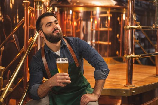 Handsome bearded male brewer resting at his brewery after working, enjoying drinking his freshly produced craft beer. Cheerful beermaker sitting near beer containers at the production factory, copy space