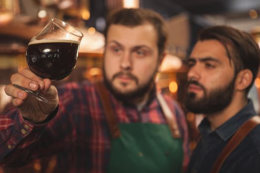 Selective focus on a glass of dark delicious beer in the hands of professional brewers. Two beermakers examining freshly brewed beer, working at their microbrewery, copy space. Business concept