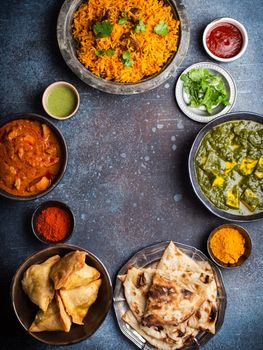 Overhead of Indian traditional dishes and appetizers: chicken curry, pilaf, naan bread, samosas, paneer, chutney on rustic background. Table with choice of food of Indian cuisine, space for text