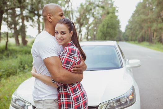 Happy mixed young couple cuddling outdoors near their car during summer road trip. Beautiful woman smiling to the camera, hugging her boyfriend while travelling together by car