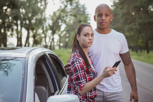 Young multiracial couple looking worried, calling roadside assistance for their broken car. Beauitful woman texting roadside help service during roadtrip with her boyfriend