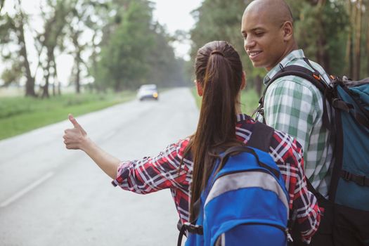 Rear view shot of a couple hitchiking on the countryside road in the forest, copy space. Cheerful handsome African man smiling at his girlfriend while backpacking together