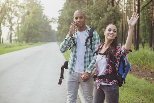Cheerful young mixed couple with backpacks hitchhiking on countryside road, copy space. Attractive young female traveler waving to stop the car, backpacking with her boyfriend
