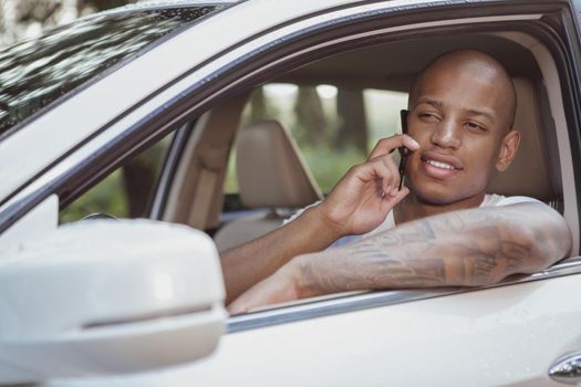 Happy handsome African man smiling, talking on the phone in his car, copy space. Attractive relaxed young man enjoying travelling by car, using mobile phone on a roadtrip