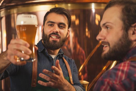 Handsome bearded brewer smiling, talking to his colleague while examining freshly brewed craft beer. Two beermakers working at microbrewery. Food and drink production, small business concept