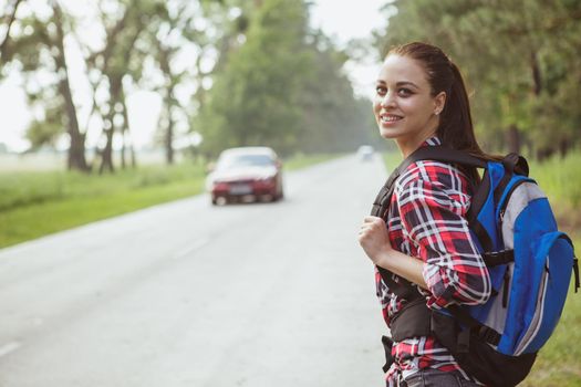 Beautiful happy young woman backpacking, smiling over her shoulder while hitchhiking on countryside road, copy space. Lovely female traveller with a backpack catching a car on the road in the woods