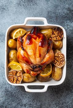 Close-up of baked or roasted whole juicy crispy organic chicken with potatoes, garlic, lemon in white casserole dish on concrete rustic background, top view