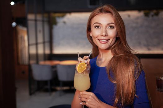 Attractive young woman smiling to the camera, holding her drink, enjoying night out at the bar. Weekend, entertainment concept