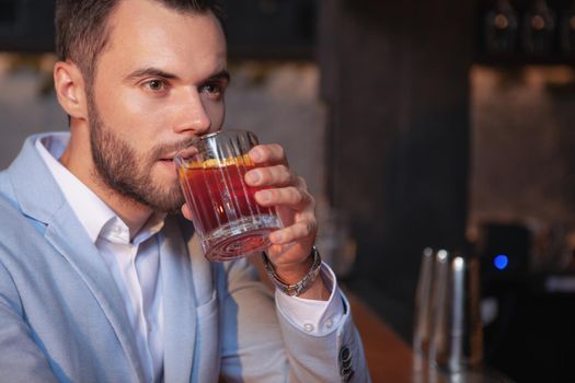 Cropped close up of a handsome bearded elegant man drinking whiskey cocktail at the bar. Attractive businessman having a drink after long working day