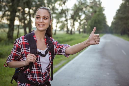 Happy young beautiful woman smiling, hitchhiking on a highway, copy space. Lovely cheerful female backpacker enjoying travelling across country, catching a car