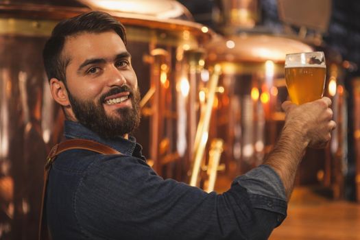 Happy bearded professional brewer smiling to the camera over his shoulder, holding glass of delicious beer, copy space. Attractive male bartender posing with a glass full of tasty beer. Bar, pub concept