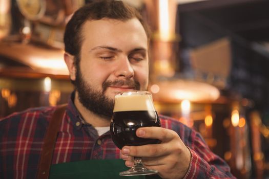 Close up of a happy male brewer smiling with his eyes closed, enjoying smelling delicious dark beer, copy space. Professional beermaker enjoying his tasty craft beer. Relax, leisure, weekend concept