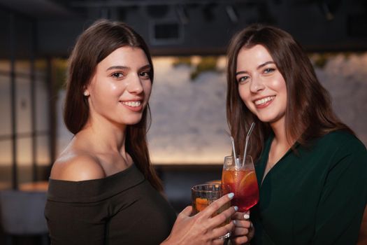 Two happy beautiful young women smiling to the camera, holding their drinks at the bar