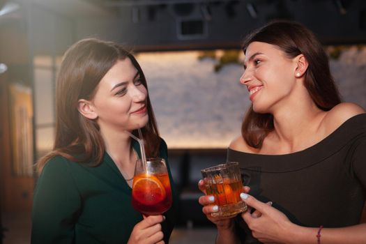 Two charming young women smiling at each other enjoying cocktails at the bar. Friendship,girlfriends concept