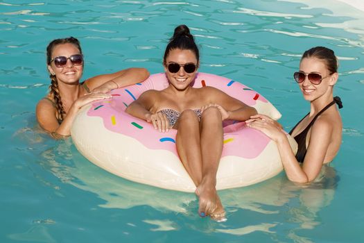Three happy beautiful female friends having a pool party, smiling to the camera, swimming on a inflatable donut together. Gorgeous women enjoying day at the pool. Recreation, travelling concept