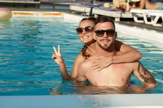 Happy young couple having fun at the swimming pool during summer vacation. Beautiful happy young woman e,bracing her handsome boyfriend while swimming in the pool together. Love, romance, tourism concept