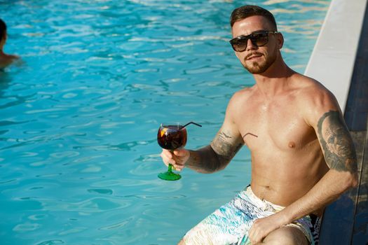 Handsome tattooed man smiling to the camera, while having a drink by the swimming pool, copy space.Attractive man smiling cheerfully having a drink near the pool. Recreation, carefree lifestyle, travel concept