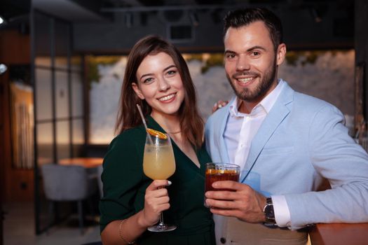 Lovely happy young couple smiling to the camera, holding their cocktails at night club. Beautiful woman on romantic date with her handsome boyfriend