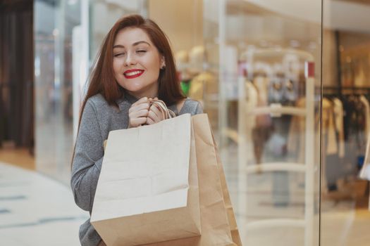 Charming young woman smiling with her eyes closed in pleasure, enjoying shopping at the mall, copy space. Attractive red lipped female customer shopping at the mall, carrying paper bags with purchase