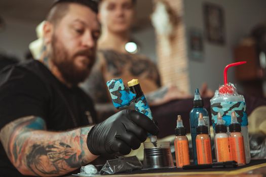 Selective focus on tattoo ink in bottles, tattooist working at his studio, copy space on the side. Young man getting new tattoo on chest. Professional tattoo artist at work. Alternative lifestyle concept