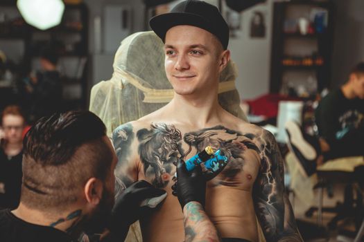 Young handsome tattooed man smiling, looking away thoughtfully while professional tattooist drawing new tattoo on his chest. Professional tattoo artist working with his client. Creativity, art concept