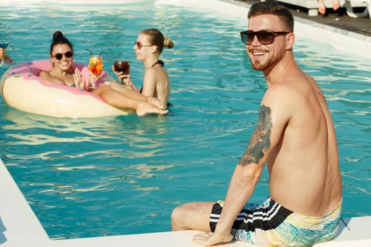 Handsome tattooed young man smiling to the camera over his shoulder, sitting on the edge of a swimming pool, his female friends having fun in the water on the background