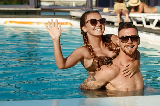 Handsome cheerful man carrying his beautiful girlfriend on his back at the swimming pool. Lovely couple having fun at the hotel poolside. Boyfriend and girlfriend travelling together in summer. Vacation concept