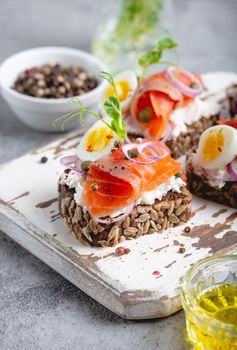 Close-up of sandwich with dark rye bread, cream cheese, salmon, onion, capers, boiled egg on white wooden cutting board, concrete background. Traditional Scandinavian open sandwich Smorrebrod.