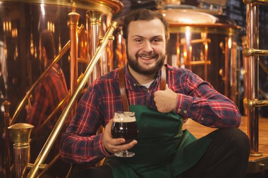 Happy young bearded brewer wearing apron sitting relaxed at his brewery, holding glass of beer showing thumbs up, copy space. Cheerful friendly beermaker enjoying craft beer at his pub