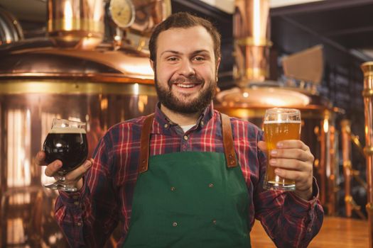 Cheerful friendly professional brewer in an apron smiling happily to the camera holding two glasses of beer, enjoying working at his microbrewery. Happy bartender serving two beers. Friendly service concept