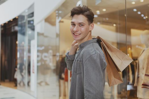 Handsome happy young man smiling to the camera over his shoulder, carrying shopping bags at the mall. Attractive male customer enjoying sale at the shopping mall. Consumerism concept