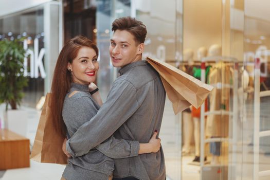 Rear view shot of a young happy couple smiling to the camera over their shoulders, carrying shopping bags at the mall, copy space. Buying goods, love concept