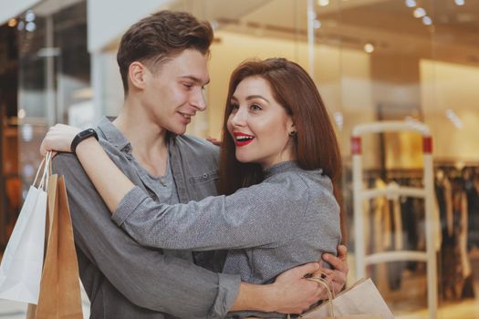 Cheerful loving young man hugging his beautiful girlfriend at the shopping mall. Young woman looking excited, while shopping at the mall with her boyfriend, copy space
