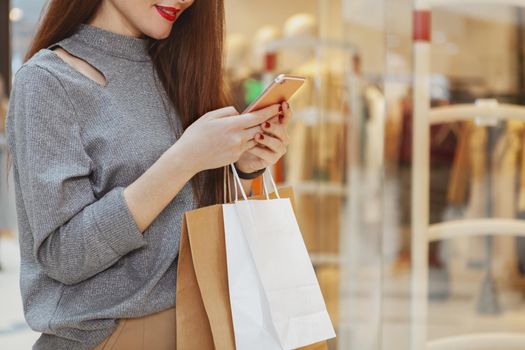 Cropped shot of a red lipped woman smiling, using smart phone while shopping at the mall. Female customer with shopping bags browsing online on her phone, copy space