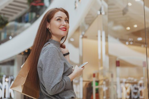 Low angle shot of a young beautiful happy woman, smiling, enjoying shopping at the mall, using her smart phone, copy space. Female customer browsing on her phone while shopping