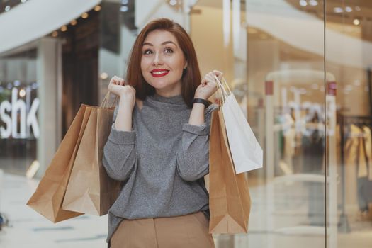 Young beautiful happy woman smiling joyfully, holding her shopping bags, enjoying sale at the mall, copy space. Excited attractive female cusotmer at the shopping mall. Consumerism, retail concept