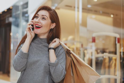 Gorgeous happy healthy woman talking on her smart phone while walking at the shopping mall, carrying paperpurchase bags. Attractive joyful female customer enjoying shopping spree at the mall