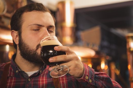 Close up of a brewer sipping delicious beer with his eyes closed, copy space. Professional beermaker enjoying working at his brewery tasting dark beer. Manufacturing, production, sales concept